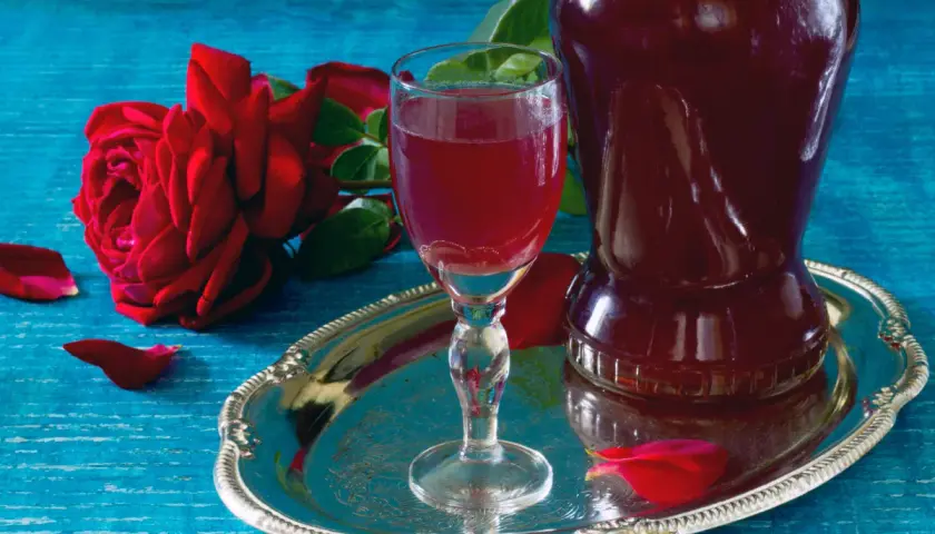 Aromatic nalewka z płatków róży w kieliszku i karafce, obok czerwonej róży i płatków na srebrnej tacy.