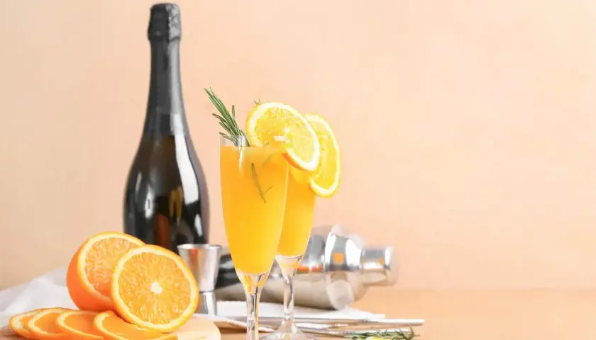 Dwa kieliszki orzeźwiającej mimosa z plasterkami pomarańczy i gałązką rozmarynu, obok butelki szampana i shakera.