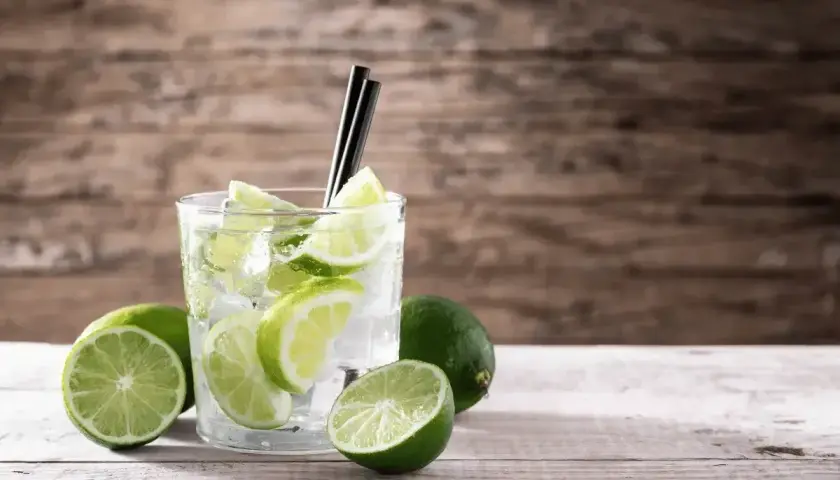 Szklanka z drinkiem, lodem i plasterkami limonki, obok kilka całych limonek.