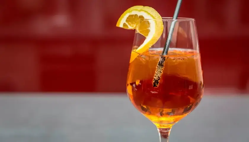 Szklanka Aperol Spritz z plasterkiem pomarańczy i metalową słomką na czerwonym tle.