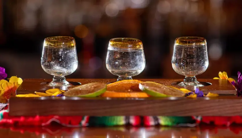 Trzy kieliszki z mezcalem, plasterki limonki i pomarańczy na drewnianej desce.