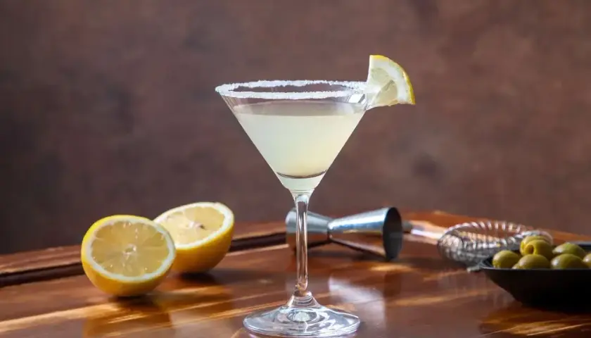 Koktajl Martini Bianco w kieliszku z solą na brzegu, udekorowany plasterkiem cytryny, obok cytryny i oliwki.