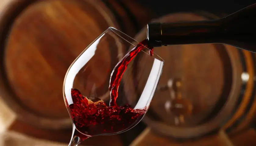 Czerwone wino, bogate w taniny, leje się z butelki do kieliszka, na tle drewnianych beczek.