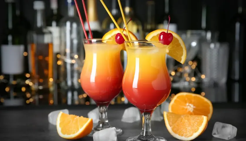 Dwa drinki z pomarańczą i wiśnią, na tle butelek i światełek.