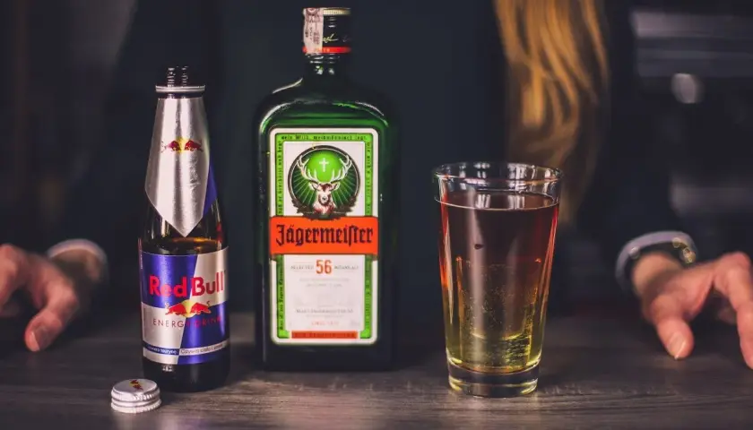 Butelka Jägermeister, puszka Red Bull i szklanka z drinkiem na drewnianym blacie.