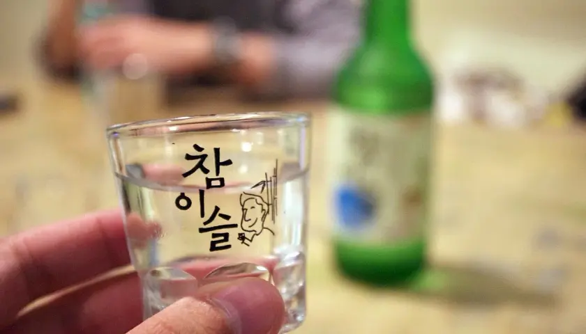 Szklanka z soju z koreańskim napisem i rysunkiem, w tle zielona butelka.