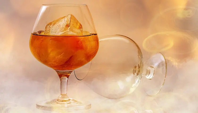 Szklanka z bursztynową brandy i lodem, obok pusta, przewrócona kieliszka.