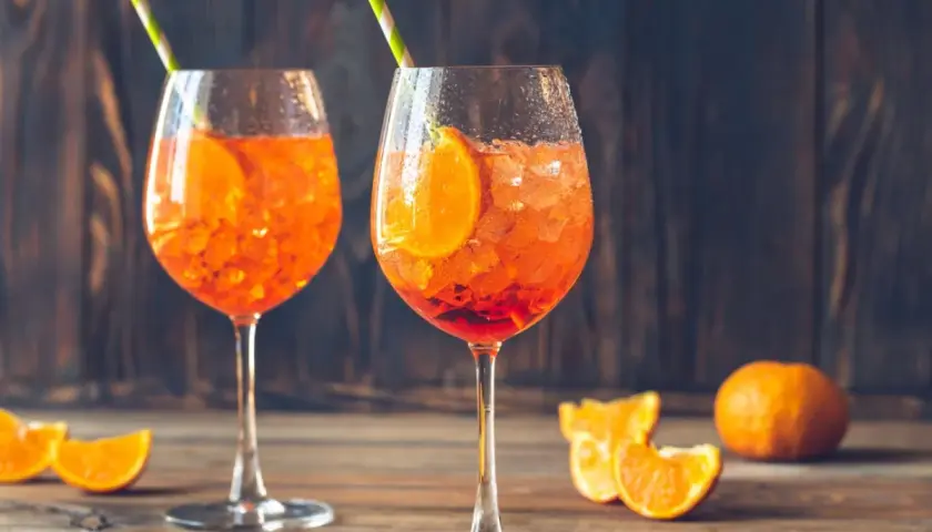 Dwa kieliszki Aperol Spritz z lodem i plasterkami pomarańczy na drewnianym stole.