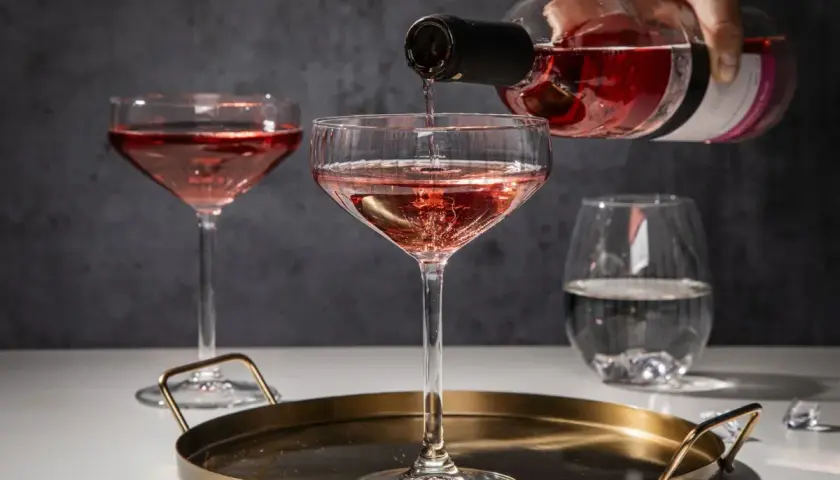 Różowe prosecco nalewane do kieliszka na tacy, obok inne kieliszki i szklanka z wodą.