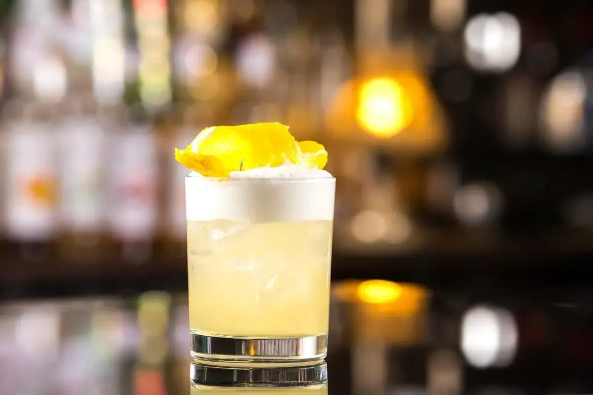 Whisky Sour z pianką i skórką cytryny w szklance na tle rozmytego baru.