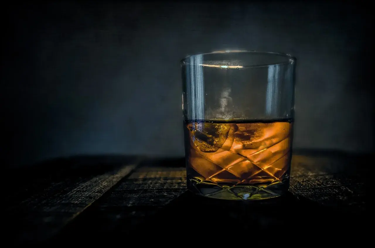 Szklanka bursztynowego trunku z lodem na drewnianym blacie. Z czego robi się whisky? Z ziaren zbóż, wody i drożdży, a potem leżakuje w beczkach.
