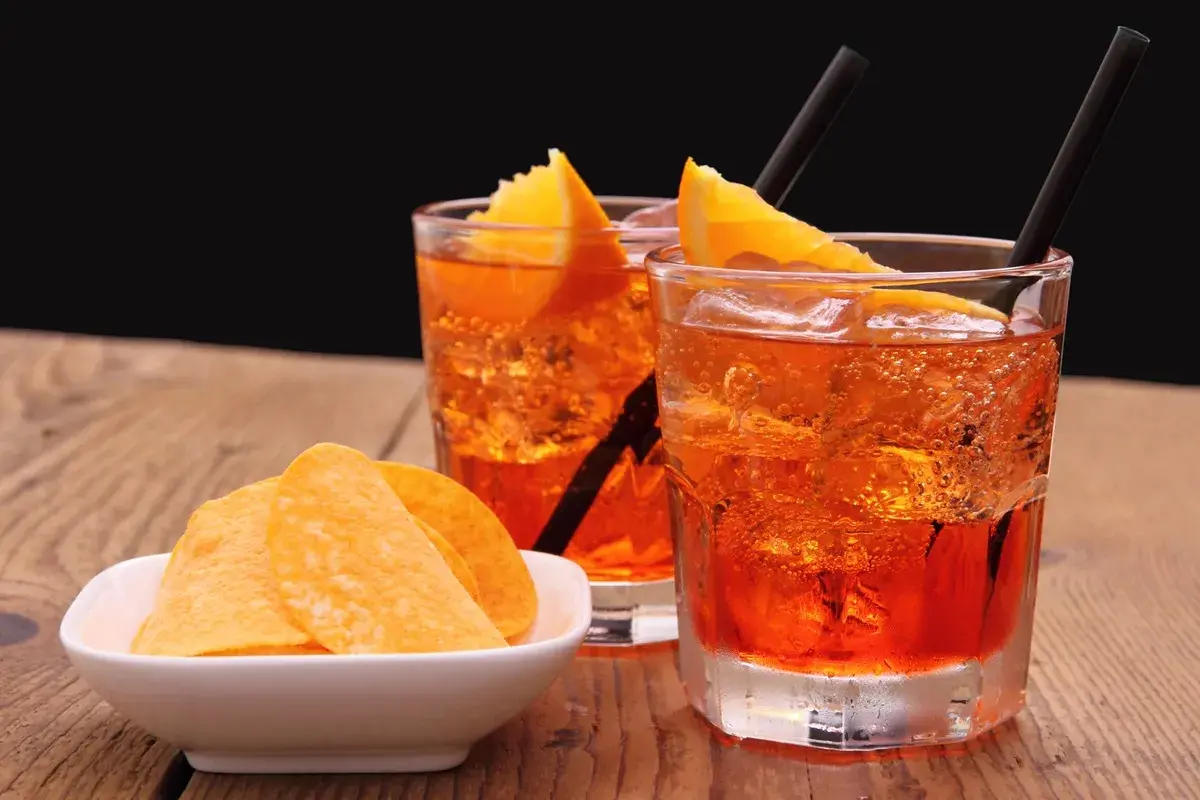 Dwa orzeźwiające drinki z pomarańczą i słomkami, obok miseczka chipsów. Idealny aperitif na letnie popołudnie.