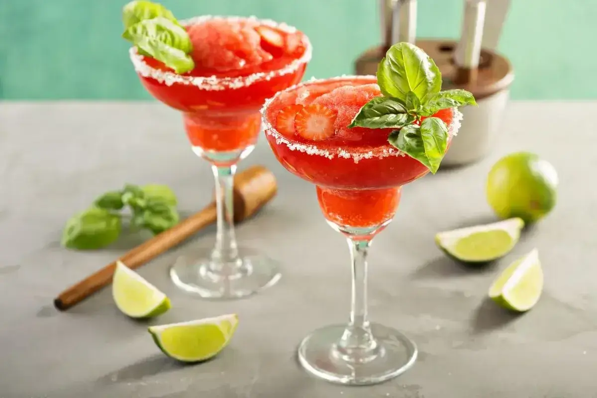 Dwa mrożone daiquiri truskawkowe w kieliszkach z cukrowym rantem, ozdobione bazylią i truskawkami.