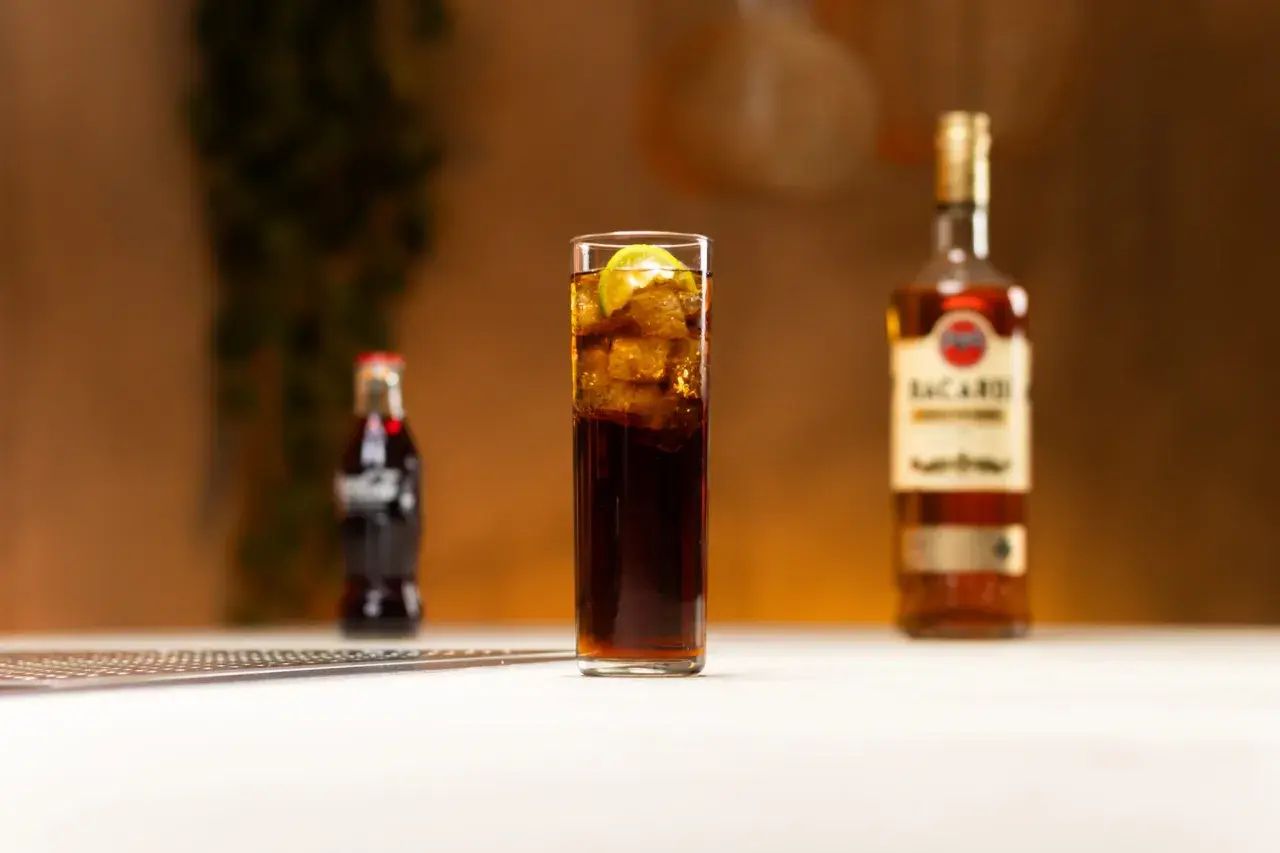 Szklanka z drinkiem z rumem Bacardi, lodem i limonką, obok butelka Coca-Coli i rumu.