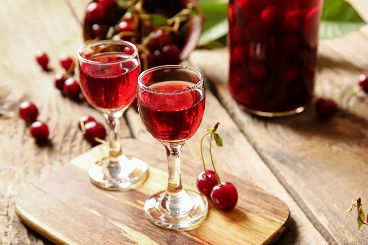 Dwa kieliszki z rubinowym winem z czereśni, obok świeże owoce i słoik pełen wiśni.