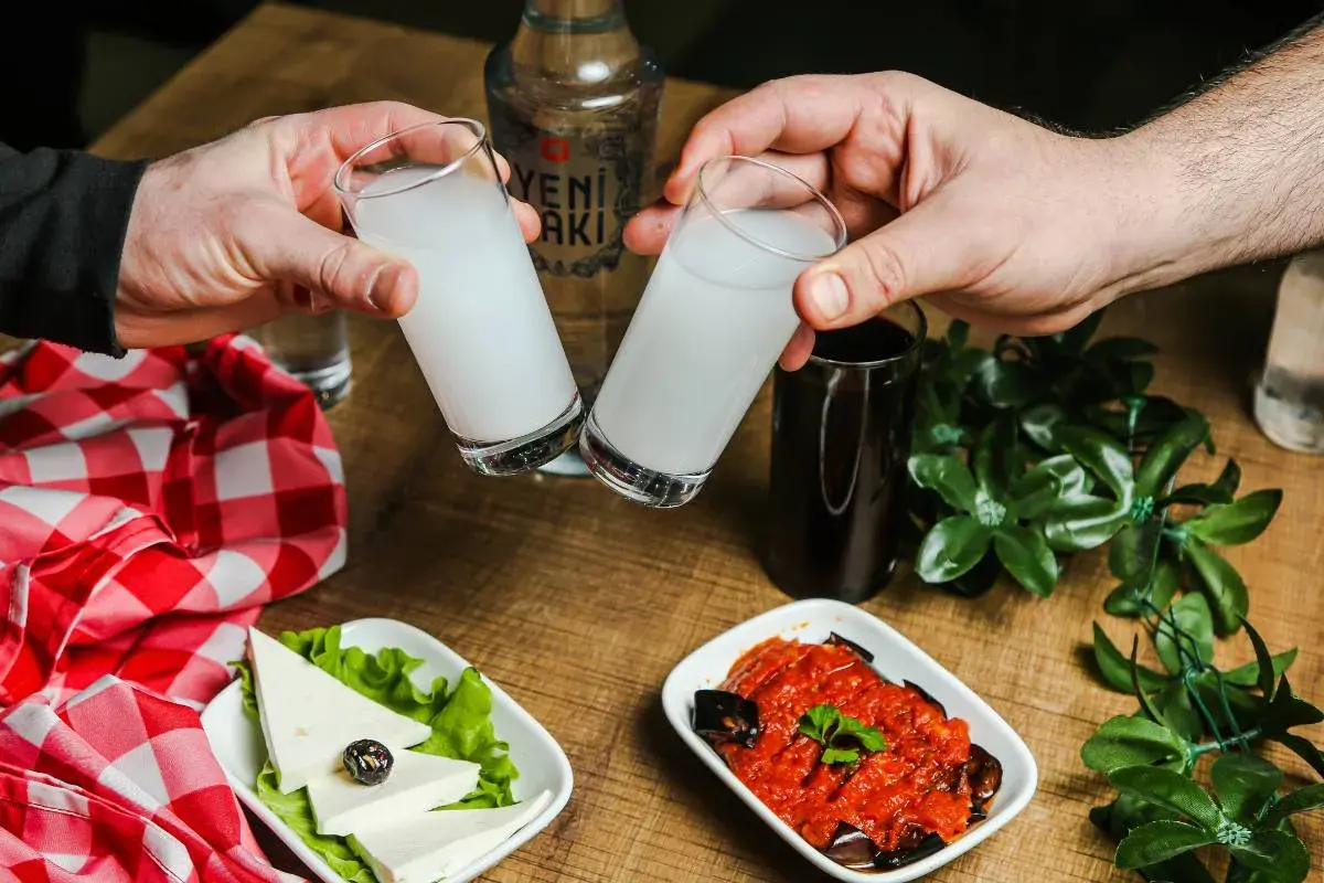 Dwie szklanki z białym alkoholem, raki, unoszą się do toastu. Na stole ser, sałatka i butelka Yeni Raki.