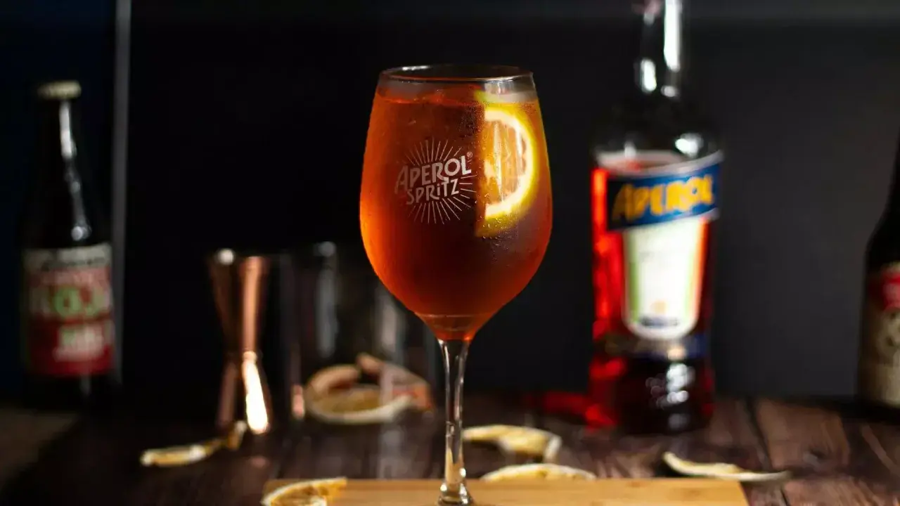 Aperol Spritz w kieliszku z plasterkiem pomarańczy, obok butelka Aperol i inne dodatki.