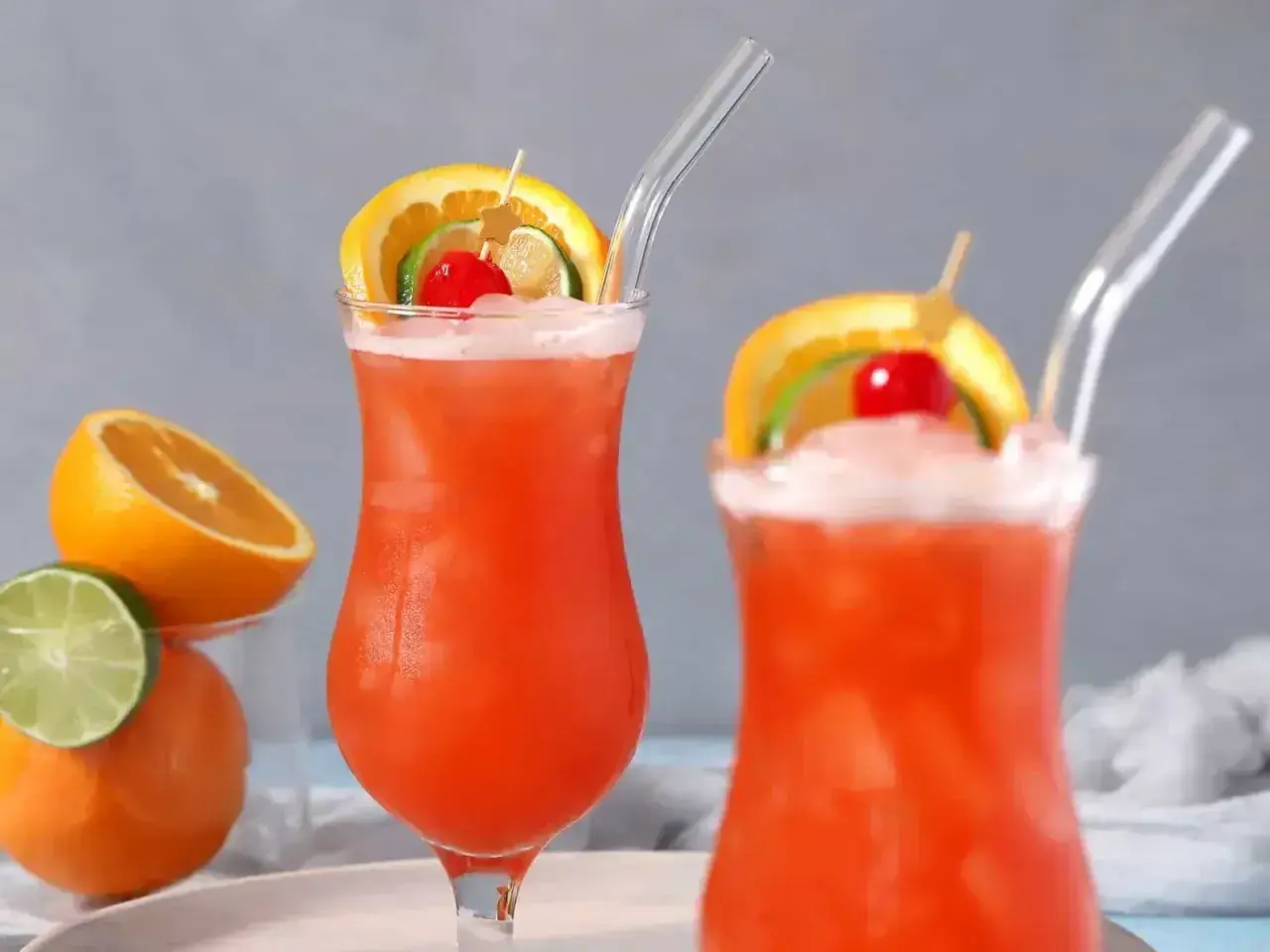 Dwa drinki z rumem, ozdobione plasterkami pomarańczy, limonki i wiśnią, z owocami w tle.