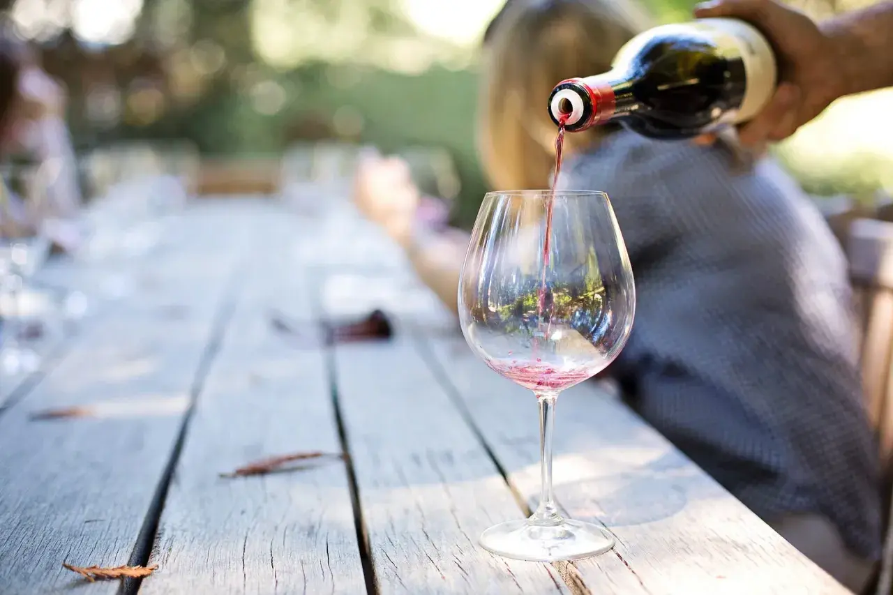 Czerwone wino Carlo Rossi nalewane do kieliszka na drewnianym stole, w tle rozmyte postacie i zieleń.
