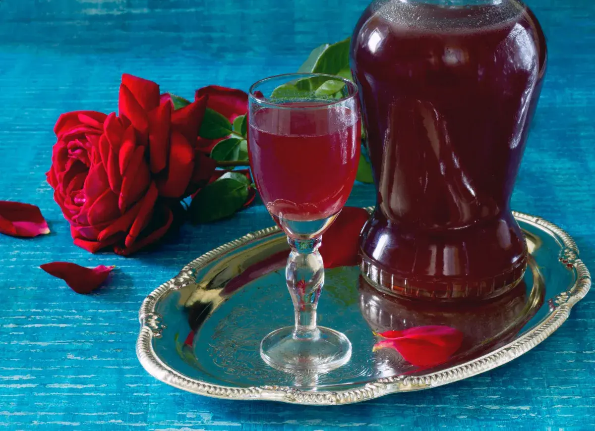 Aromatic nalewka z płatków róży w kieliszku i karafce, obok czerwonej róży i płatków na srebrnej tacy.