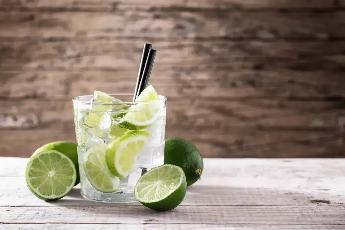 Szklanka z drinkiem, lodem i plasterkami limonki, obok kilka całych limonek.