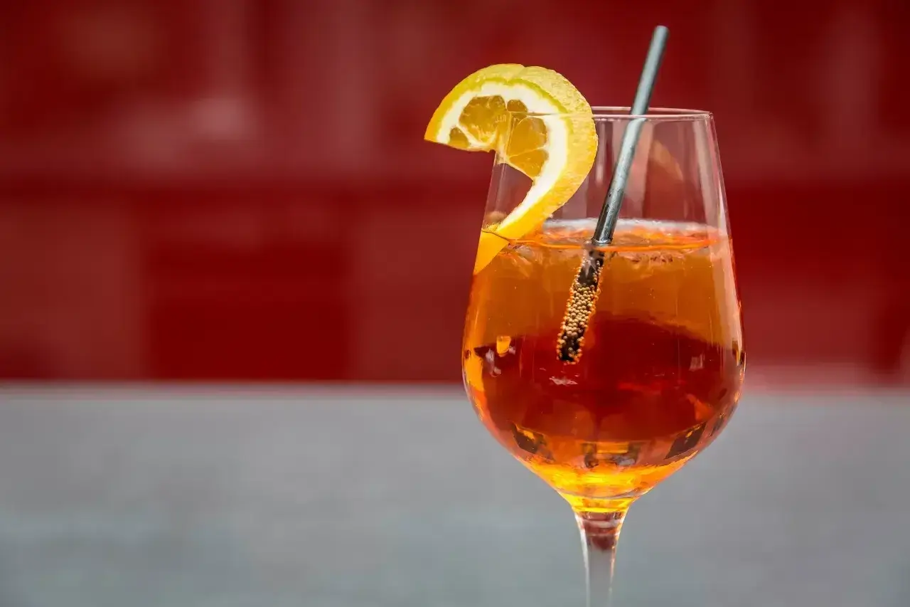 Szklanka Aperol Spritz z plasterkiem pomarańczy i metalową słomką na czerwonym tle.