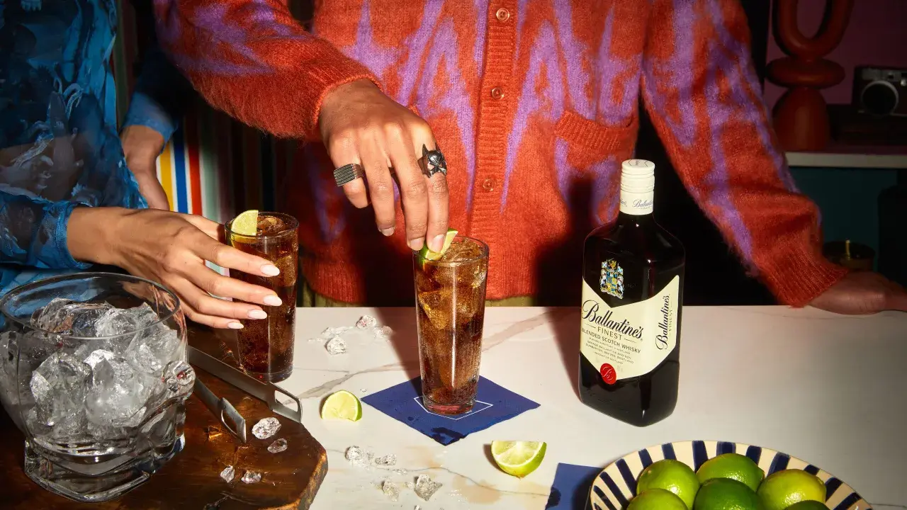 Dwie osoby przygotowują drinki z whisky i colą, z butelką Ballantine's, lodem i limonkami.