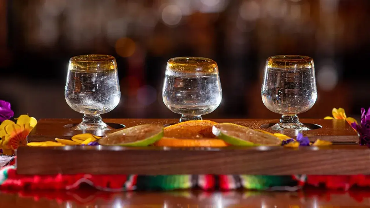 Trzy kieliszki z mezcalem, plasterki limonki i pomarańczy na drewnianej desce.