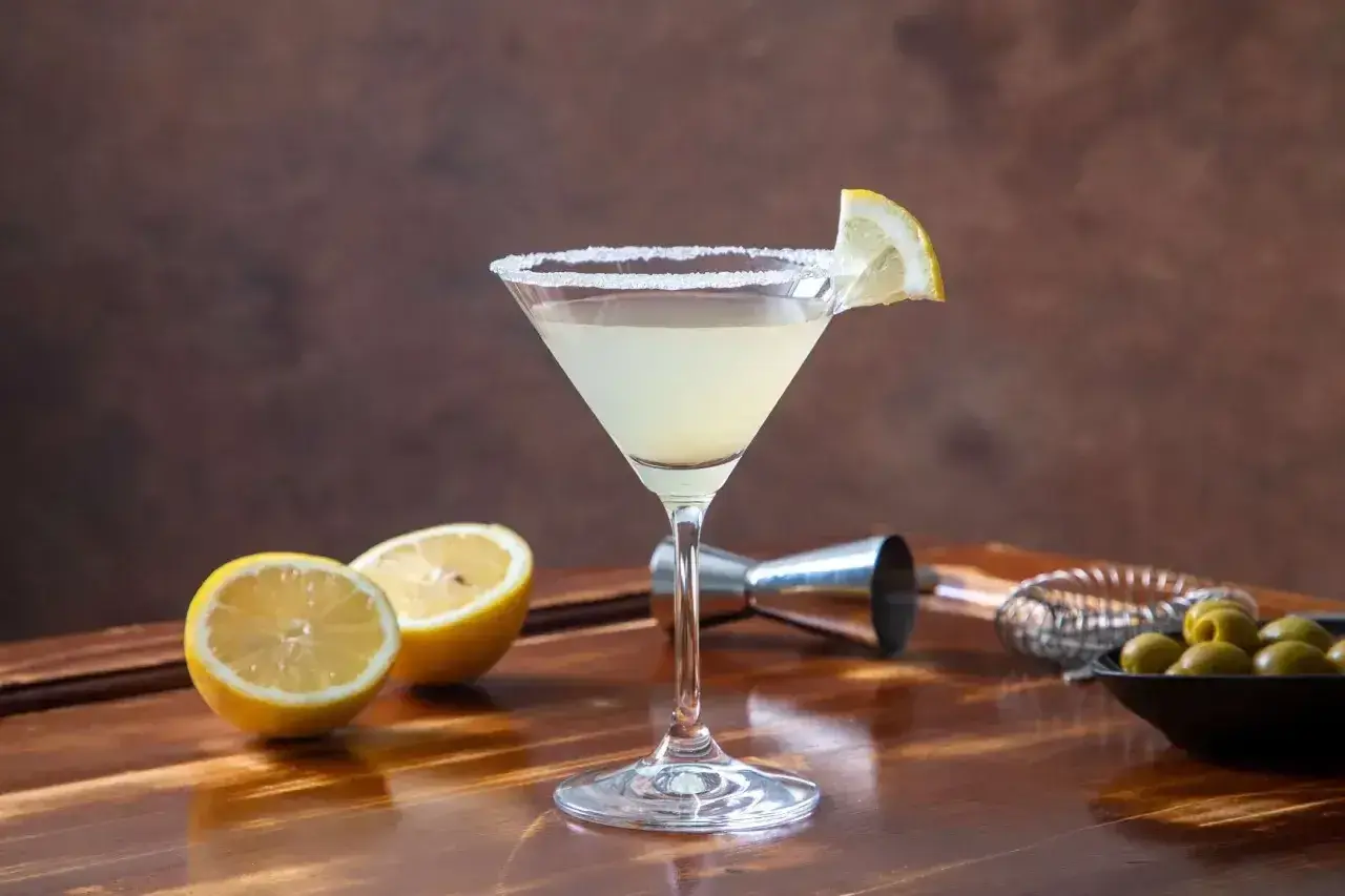 Koktajl Martini Bianco w kieliszku z solą na brzegu, udekorowany plasterkiem cytryny, obok cytryny i oliwki.
