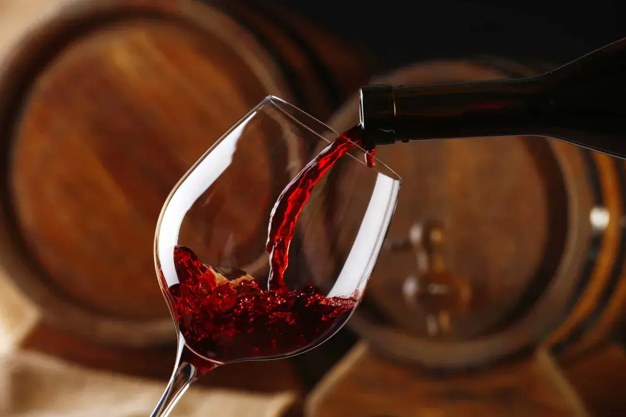 Czerwone wino, bogate w taniny, leje się z butelki do kieliszka, na tle drewnianych beczek.