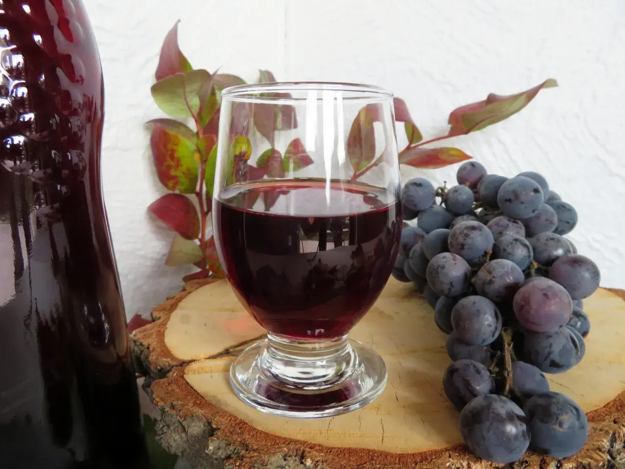 Domowy przepis na wino z winogron 10l. Kieliszek czerwonego wina, butelka, kiść ciemnych winogron i gałązka z liśćmi.