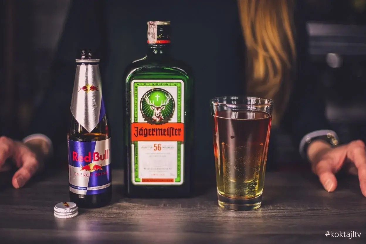 Butelka Jägermeister, puszka Red Bull i szklanka z drinkiem na drewnianym blacie.