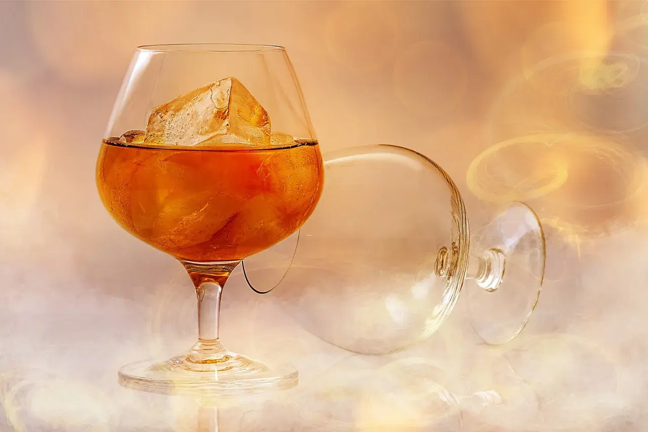Szklanka z bursztynową brandy i lodem, obok pusta, przewrócona kieliszka.