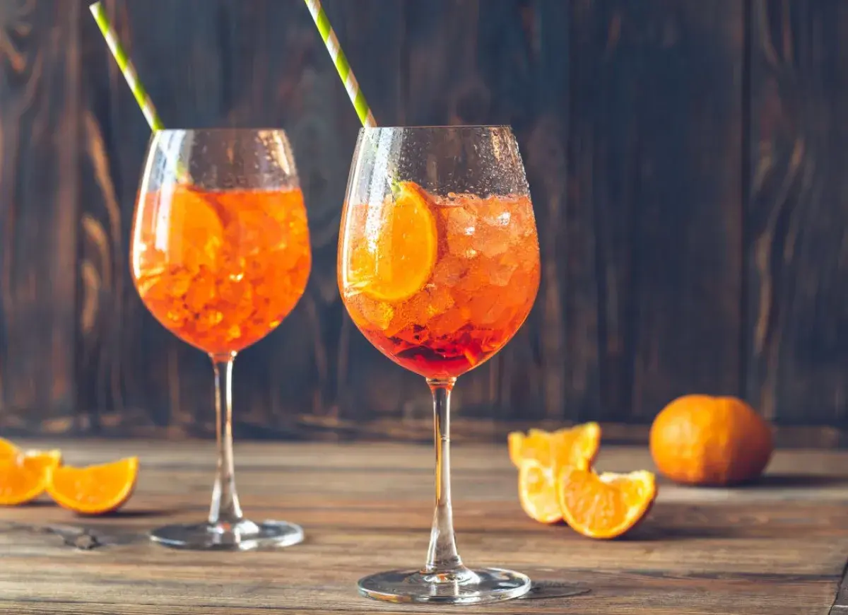 Dwa kieliszki Aperol Spritz z lodem i plasterkami pomarańczy na drewnianym stole.