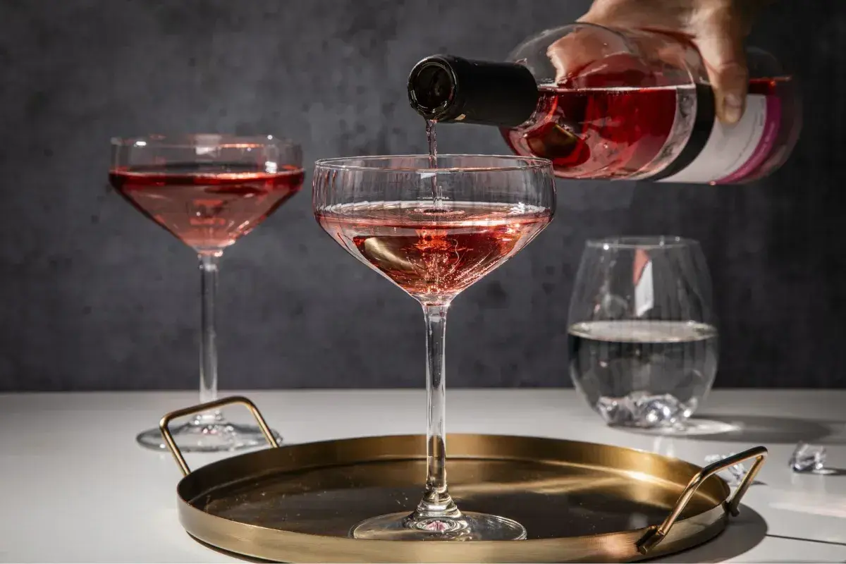 Różowe prosecco nalewane do kieliszka na tacy, obok inne kieliszki i szklanka z wodą.