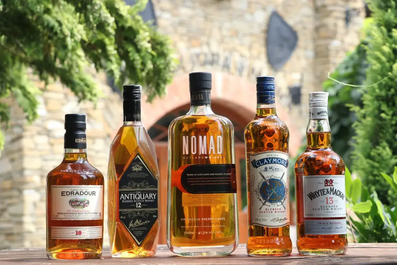 Pięć butelek różnych rodzajów whisky: Edradour, Antiquary, Nomad, Claymore i Whyte & Mackay, stojących na drewnianym stole.