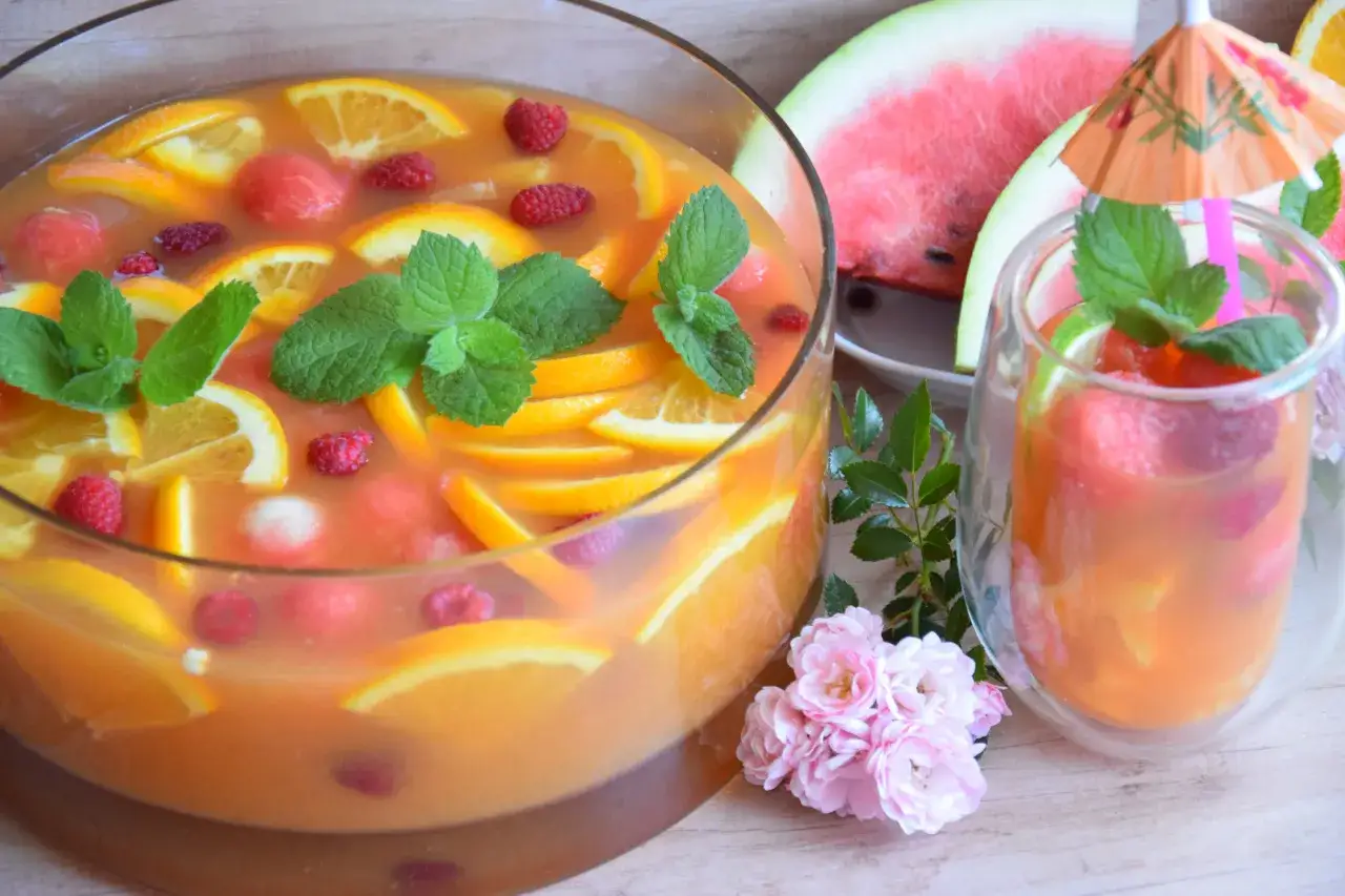 Orzeźwiający jungle juice w dużej misie, pełen plastrów pomarańczy, malin i kawałków arbuza. Obok kieliszek z tym samym napojem i parasolką.