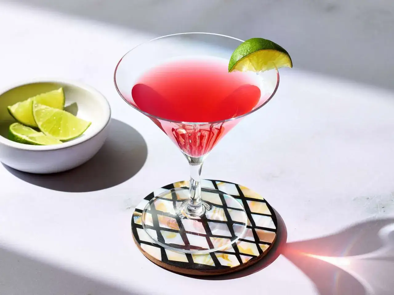 Elegancki cosmopolitan z limonką, obok miseczka z ćwiartkami limonki, na ozdobnej podstawce.