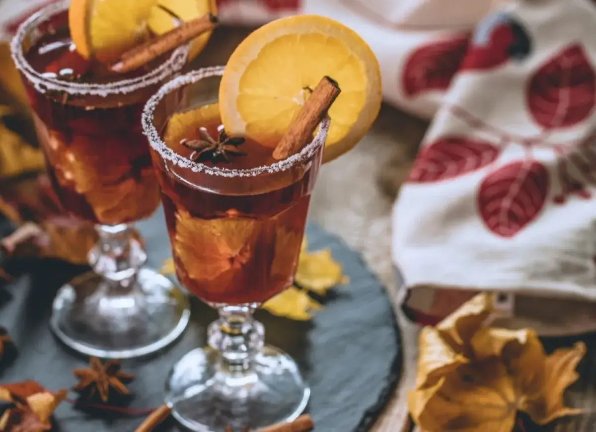 Dwa kieliszki z rozgrzewającym krupnikiem alkoholowym, ozdobione plastrami pomarańczy i laskami cynamonu, na tle jesiennych liści.