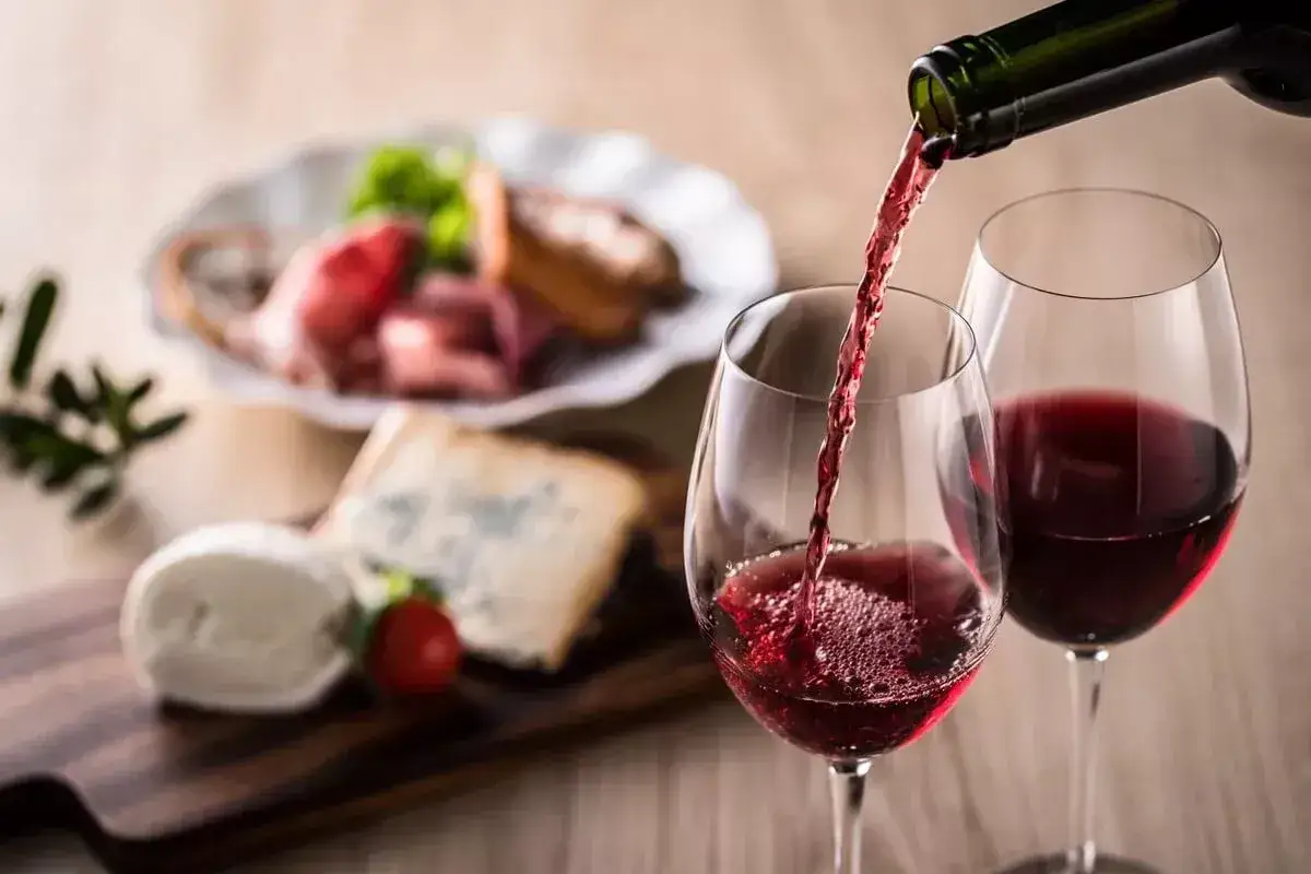 Czerwone wino, być może słodkie, nalewane do kieliszka, obok deska serów i wędlin.