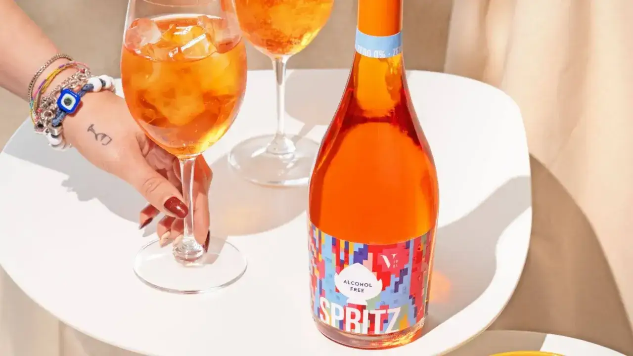 Ręka z bransoletkami sięga po kieliszek z orzeźwiającym aperol bezalkoholowy. Obok butelka z kolorową etykietą.