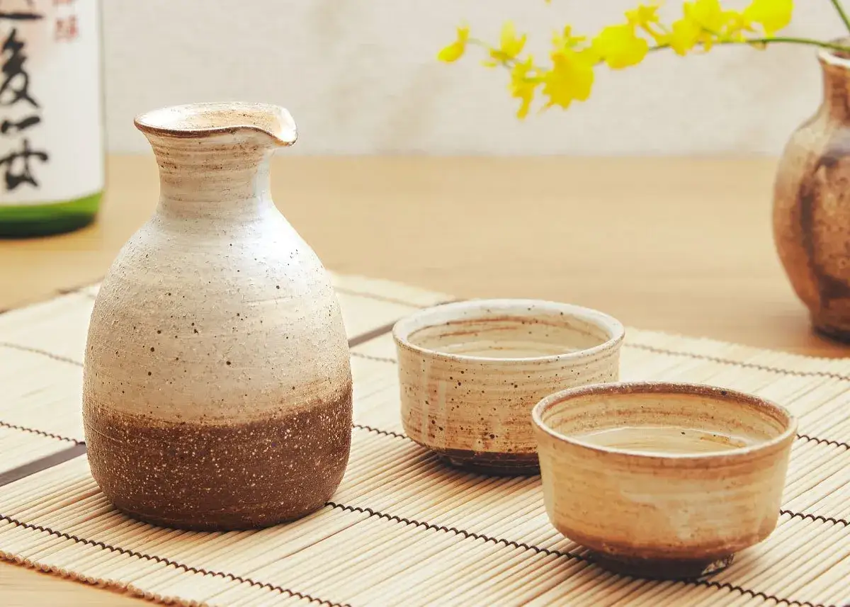 Ceramiczny dzbanuszek i dwie czarki do sake, ozdobione żółtymi kwiatami.