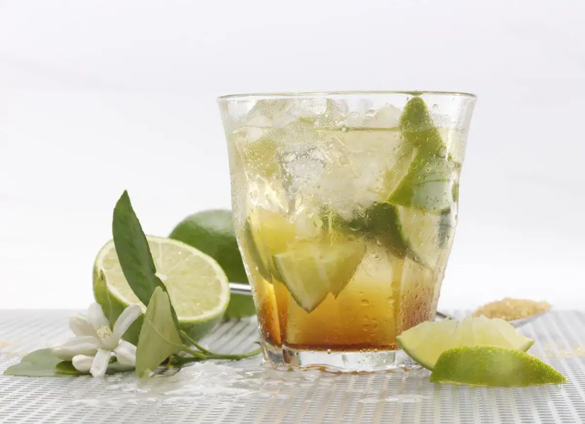 Orzeźwiająca caipirinha przepis: drink z lodem, limonką i cukrem. Obok limonki i kwiaty.