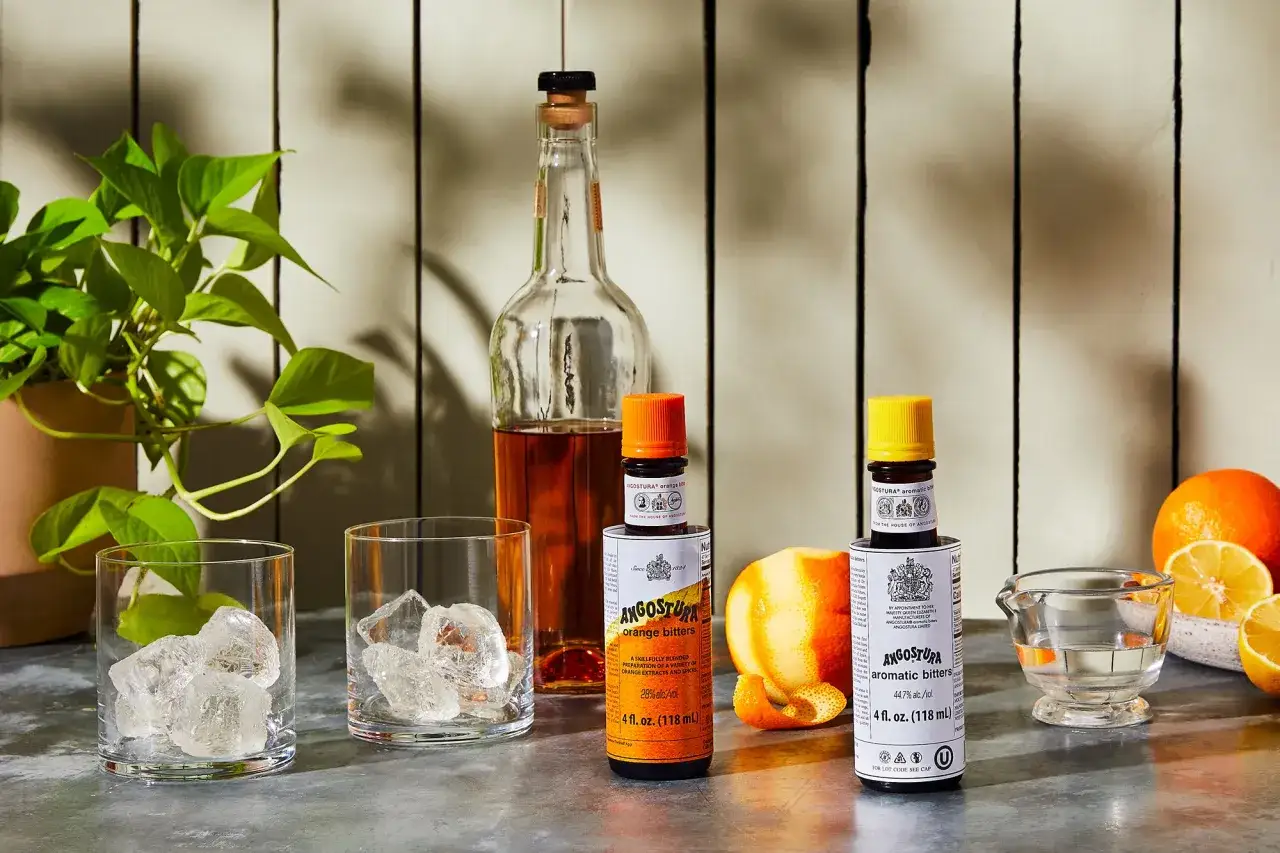 Dwie buteleczki Angostura bitters (pomarańczowe i aromatyczne) obok szklanek z lodem i owoców. Idealne do drinków, jeśli zastanawiasz się, angostura co to.