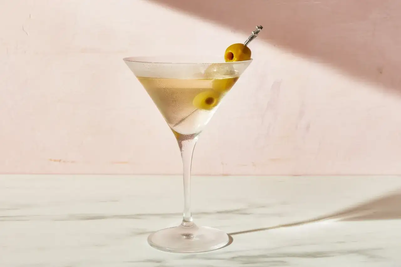 Elegancki, schłodzony dirty martini z dwiema oliwkami na wykałaczce, w kieliszku na tle różowej ściany.
